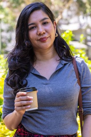 Young woman smiling and holding coffee cup in sunlit courtyard wearing gray top