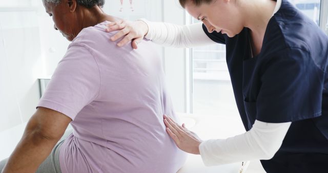 Female physiotherapist gently assessing elderly woman's back pain in bright clinical setting. Ideal for promoting physiotherapy clinics, healthcare services, elderly care, rehabilitation centers, and ads focused on medical treatments for back problems.