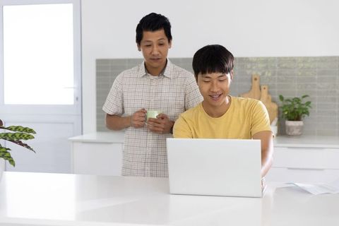 Asian father watching son using laptop at bright modern kitchen island for work from home lifestyle