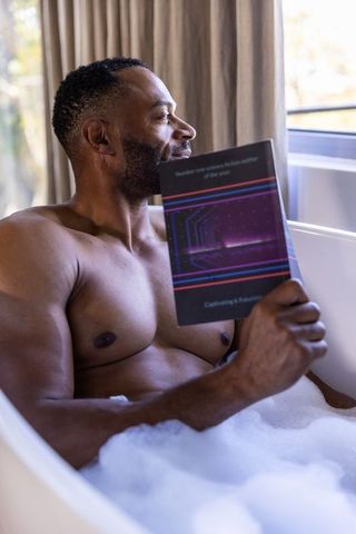 African american man relaxing in bubble bath reading paperback near window