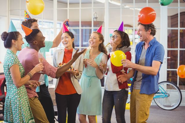 Diverse group of business people celebrating a birthday in the office. Colleagues are wearing party hats, holding balloons, and enjoying drinks and cake. Ideal for showcasing workplace culture, team bonding, and festive office environments.