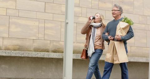 Mature couple walking with groceries and coffee in urban neighborhood on overcast day