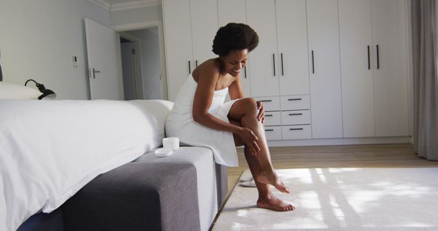 Happy african american woman apply cream on legs in bedroom. domestic lifestyle, spending free time at home.