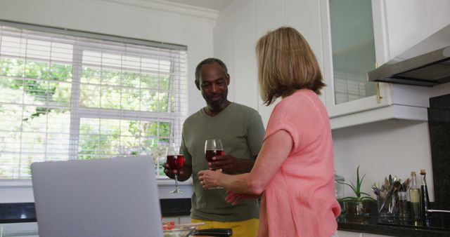 Diverse senior couple in kitchen using laptop and drinking wine. staying at home in isolation during quarantine lockdown.
