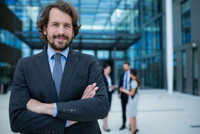 Businessman standing with arms crossed and smiling in front of a modern office building. Colleagues are seen in the background, engaged in conversation. Ideal for use in corporate websites, business presentations, leadership articles, and professional networking profiles.