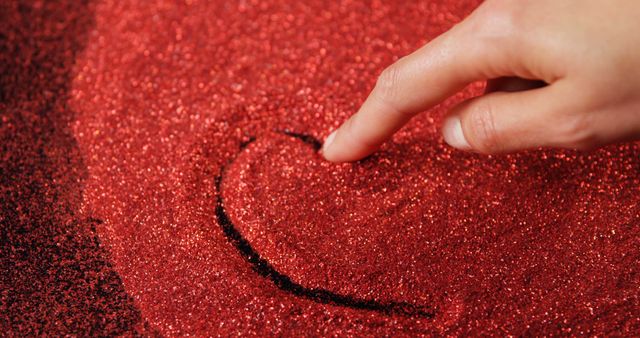Close-up view of a hand drawing a heart shape in a pile of red glitter sand. Great for Valentine's Day projects, advertisements promoting love and affection, creative art concepts, and romance-themed blog posts.