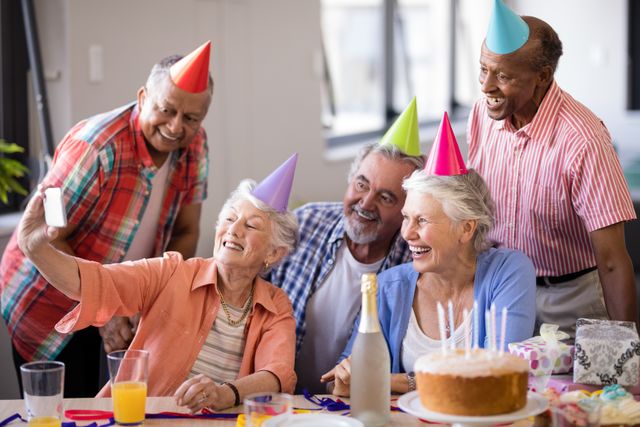 Senior friends celebrating a birthday, taking a selfie with a mobile phone. They are wearing party hats and smiling around a table with a birthday cake and drinks. Perfect for themes of friendship, aging, celebrations, and social gatherings.