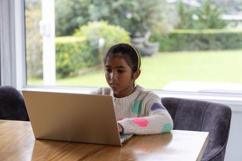 Asian school-age girl studying on laptop at dining table near window for remote learning
