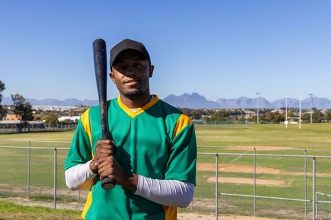 Confident African American Baseball Player Holding Bat on Sunlit Field with Mountain View