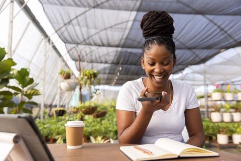 Smiling african american woman voice-recording on smartphone while journaling in greenhouse