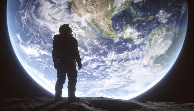 Astronaut standing on moon ridge viewing massive earthrise over rocky horizon silhouette backlit by