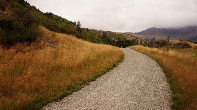 Winding gravel path surrounded by tall grassy hills leading towards distant mountains. This peaceful scene is perfect for nature presentations, travel blogs, outdoor adventure ads, and rural tourism campaigns. Ideal background for quotes and motivational content.