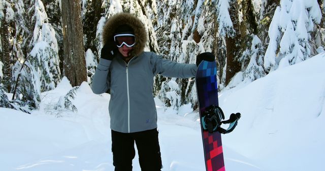Person standing in a snowy forest adjusting goggles, holding a snowboard with vibrant design. Ideal for outdoor winter sports, adventure travel, and winter sports apparel campaigns.