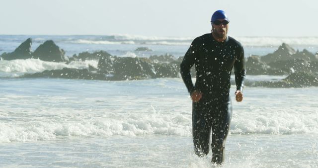 Man wearing wetsuit, swim cap, and goggles runs out of ocean. Waves crash on shore. Suitable for content about triathlons, endurance events, athletic training, fitness lifestyles or promoting sporting goods.