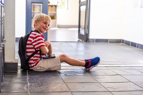 Smiling schoolboy sitting in hallway with backpack holding smartphone and wearing sneakers
