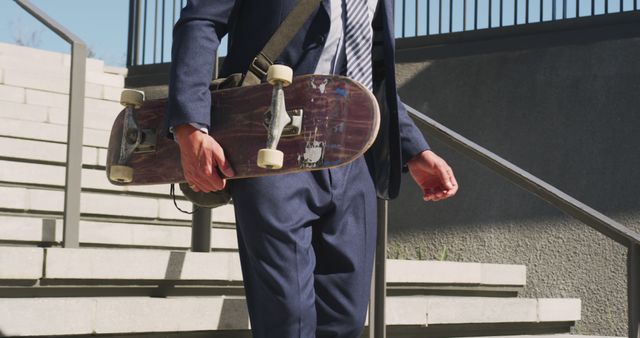 Business professional in a suit carrying a skateboard while descending outdoor stairs on a sunny day. Suitable for themes of urban lifestyle, modern business culture, unconventional work environments, and balancing work and play.