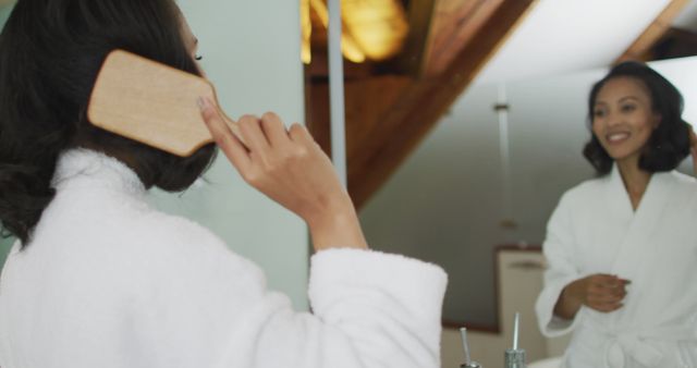 Biracial woman wearing bathrobe looking at mirror and brushing her hair. domestic life, spending quality free time relaxing at home.