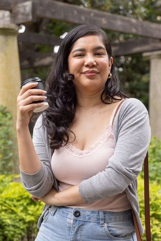 Young woman standing in garden holding takeaway coffee cup under wooden pergola