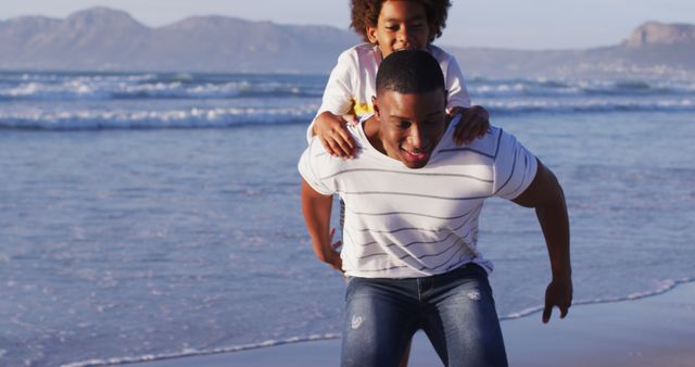 Father playing with his daughter at the beach during sunset. Background shows scenic ocean and mountains. Great for themes involving family bonding, playful activities, nature outings, and happy childhood moments.