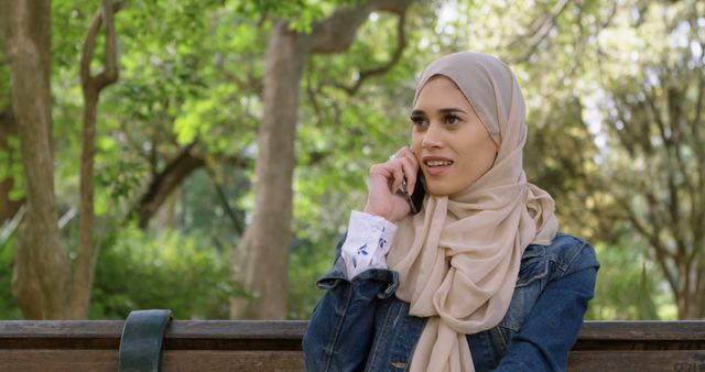 Muslim woman is engaged in a phone conversation while sitting on a park bench. She is wearing a hijab and looks happy and relaxed. This image can be used for topics related to communication, technology, relaxation, outdoor activities, cultural diversity, and lifestyle.