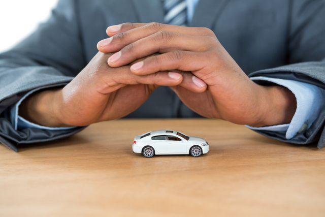 Businessman covering miniature car with hands symbolizes concepts of security, insurance, and protection. Ideal for content related to auto insurance, business investments, vehicle safety, and corporate responsibility. Suitable for websites, blogs, and promotional materials that address business or automotive services.
