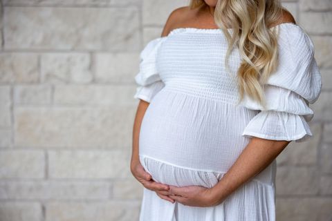 Pregnant woman holding belly in white dress against brick wall