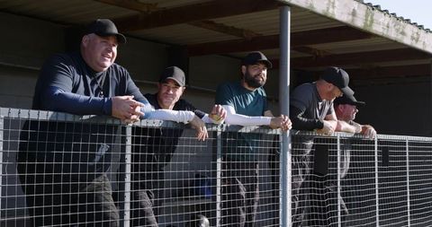 Team members leaning on dugout railing watching softball holding yellow ball and smiling