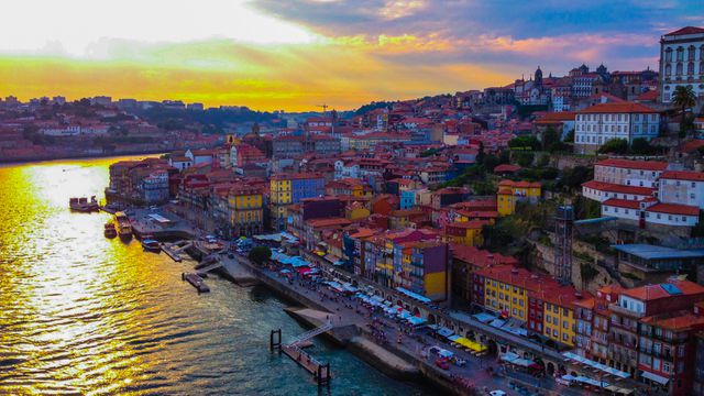 Aerial view capturing the vibrant and colorful buildings along the riverside of Porto, Portugal at sunset. The Douro River glistens as the sun sets, enhancing the rich architectural beauty of the city. Ideal for use in travel promotions, tourism brochures, postcards, and social media campaigns highlighting European destinations.