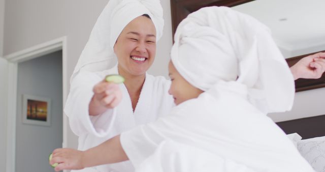 Mother and daughter spend quality time together enjoying a spa day at home. They are dressed in white bathrobes and have towels wrapped around their heads, making use of cucumbers for a refreshing skincare routine. This image can be used to depict family bonding, wellness, self-care routines, and relaxing activities suited for advertisement in beauty, wellness, or family-oriented products.