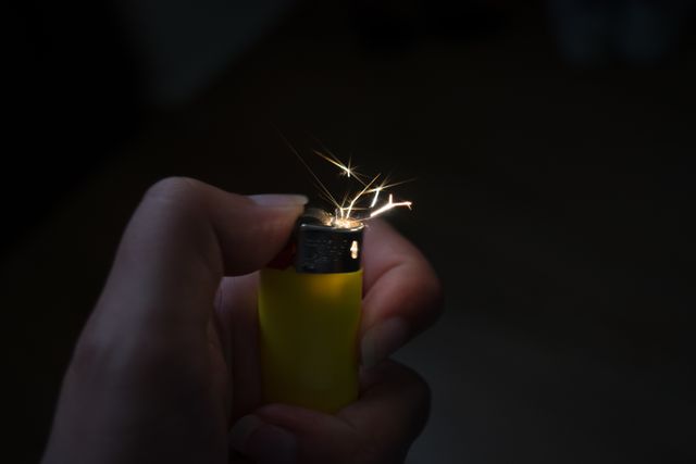 Hand holding and lighting a yellow lighter with visible sparks. Useful for content on fire safety, ignition mechanisms, DIY projects, or demonstrating lighter functionality in instructional materials.