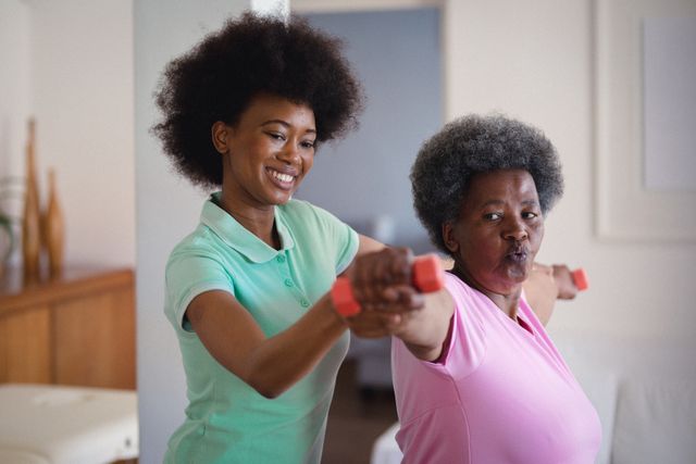 African American physiotherapist assisting senior woman with dumbbell exercises at home. Ideal for use in articles or advertisements related to senior healthcare, home fitness, physical therapy, elderly wellness, and active aging. Can be used to promote home care services, fitness programs for seniors, or physiotherapy practices.