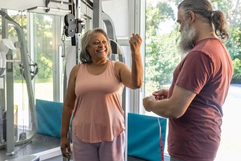 Senior african american woman flexing during fitness assessment with trainer in home gym