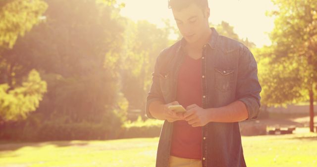 Young man texting in sunlight in park. Perfect for technology, communication, and outdoor-themed content.