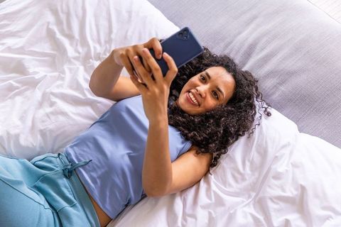 African american woman relaxing on bed using smartphone smiling in cozy bedroom lifestyle