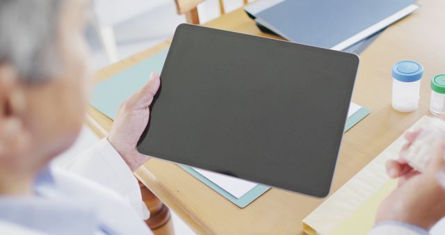 Elderly woman is holding a digital tablet in a professional office environment with folders and documents on the table. Useful for themes on modern technology use by older adults, digital healthcare services, business meetings, and office work.