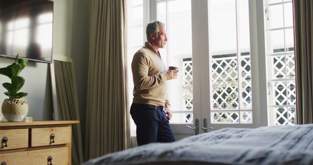 Image shows a middle-aged man wearing casual attire enjoying a morning coffee by the window in his cozy bedroom. The serene setting features sunlight coming through the large windows, highlighting the peaceful ambiance of the home interior. Ideal for use in advertisements promoting coffee brands, cozy home interiors, morning routines, or lifestyle content focusing on relaxation and home comfort.