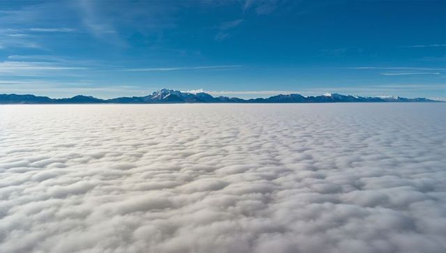 Aerial cloud deck rolling beneath snowy mountain peaks with sunlit summits and contrails