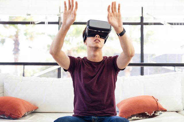 Asian teenage boy sitting on a couch at home, using a virtual reality headset and gesturing with his hands. Ideal for illustrating concepts related to modern technology, virtual reality, gaming, and immersive digital experiences. Suitable for use in articles, blogs, advertisements, and educational materials about VR technology and its applications in everyday life.