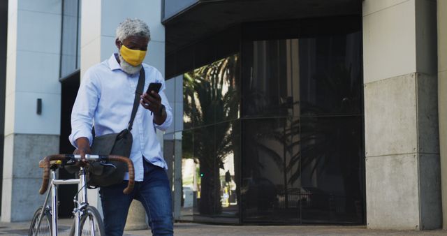 African american senior man wearing face mask with bicycle using smartphone in corporate park. hygiene and social distancing during coronavirus covid-19 pandemic.