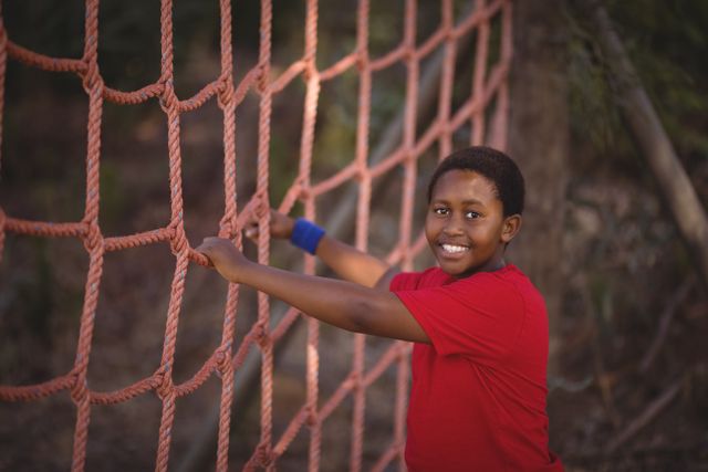 Boy smiling while climbing a net in an outdoor obstacle course. Ideal for use in content related to children's activities, outdoor adventures, summer camps, physical fitness, and teamwork exercises.