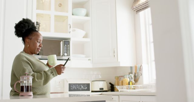 Woman standing in bright, modern kitchen holding mug of coffee while checking smartphone. Kitchen features white cabinets and contemporary design. Useful for lifestyle, technology, home environment, and relaxation themed projects.