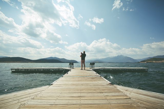 Father holding child on a wooden pier overlooking a calm lake under a partly cloudy sky, with mountains in the distance. Ideal for themes of family bonding, nature relaxation, outdoor activities, and summer vacations.