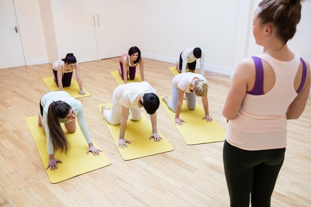 Fitness trainer guiding a group of people practicing yoga on yellow mats in a fitness studio. Ideal for use in articles or advertisements related to fitness, wellness, group exercise classes, yoga studios, and healthy living.