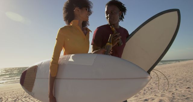 Couple carrying surfboards while enjoying beers on the beach. They engage in friendly conversation under the clear sky by the ocean. Ideal for themes of leisure, summer vacations, beach lifestyle, or active lifestyles. Perfect for use in travel brochures, lifestyle blogs, or beachwear advertisements.
