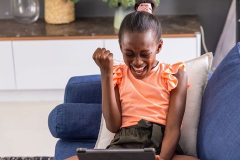 African american girl cheering on tablet sitting on blue sofa, joyful learning victory