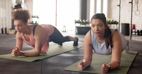 Diverse women holding forearm planks on mats with smartwatches in sunlit gym studio