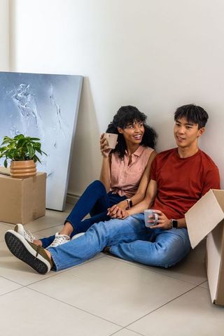 Multicultural couple sitting on floor taking break while unpacking new home with mugs