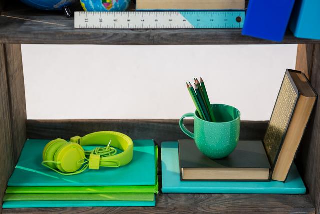 Organized shelf featuring books, green headphones, a mug with pencils, and a ruler. Ideal for use in educational content, study space inspiration, or organizational tips. Perfect for illustrating concepts of tidiness, productivity, and school or office environments.