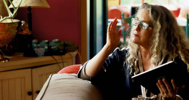 Mature woman with curly hair sitting on couch, reading book and reflecting. Home atmosphere suggesting relaxation and comfort. Useful for themes about reading, literature, senior lifestyle, relaxation, and coziness.
