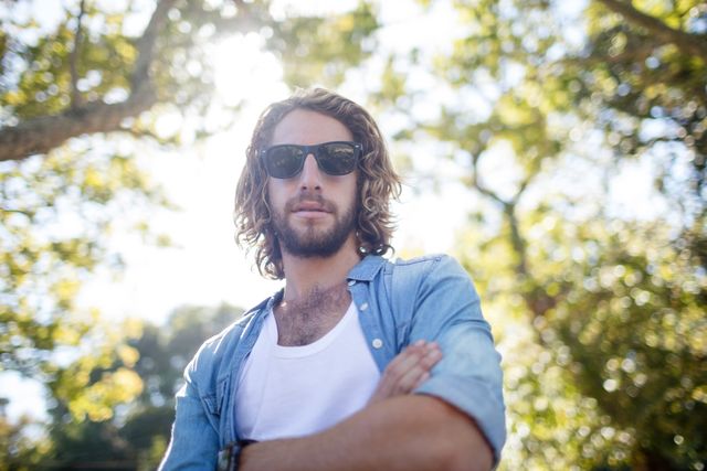 Young man standing confidently in a park with arms crossed, wearing sunglasses and casual attire. Sunlight filters through the trees, creating a relaxed and natural atmosphere. Ideal for use in lifestyle blogs, fashion articles, or advertisements promoting outdoor activities and casual wear.